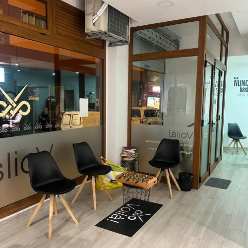 Interior de un salón de belleza con sillas negras, una mesa de futbolito y un ambiente moderno. Se puede ver un aire acondicionado y ventanas grandes que permiten la entrada de luz natural.