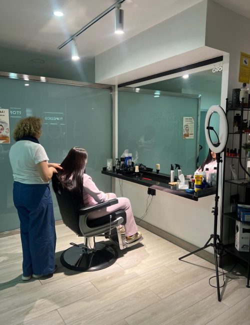 Estilista realizando un tratamiento capilar a una clienta en un salón moderno, con productos de belleza visibles en la mesa y un espejo reflejando el ambiente.